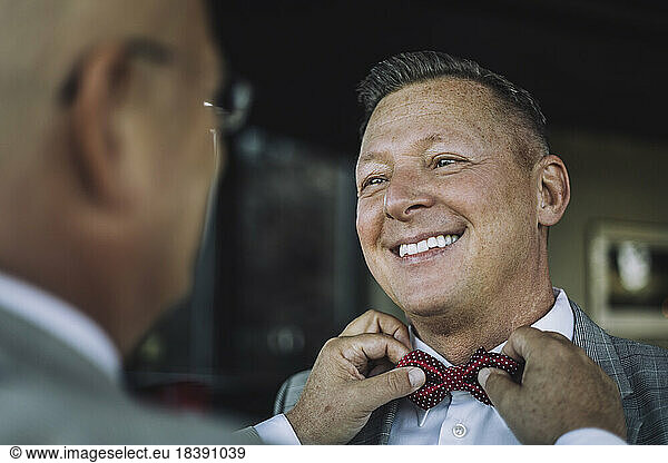 Smiling gay man looking at partner adjusting bow tie before wedding