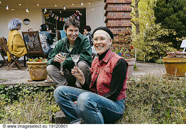 Smiling gay women sitting together holding wineglasses near plants