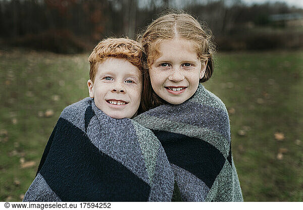 Smiling girl and brother wrapped in blanket on meadow