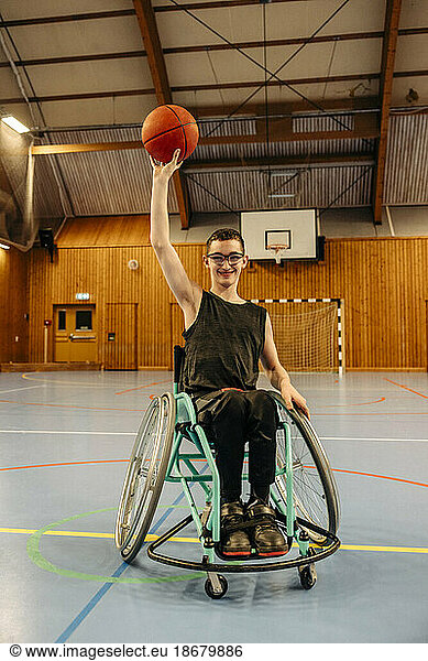 Smiling girl balancing basketball while sitting on wheelchair at sports court