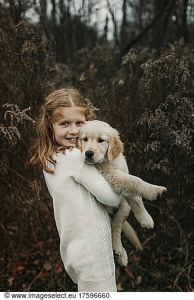 Smiling girl carrying golden retriever puppy standing in front of tree
