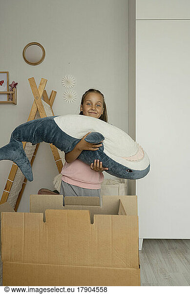 Smiling girl carrying shark toy standing by box at home
