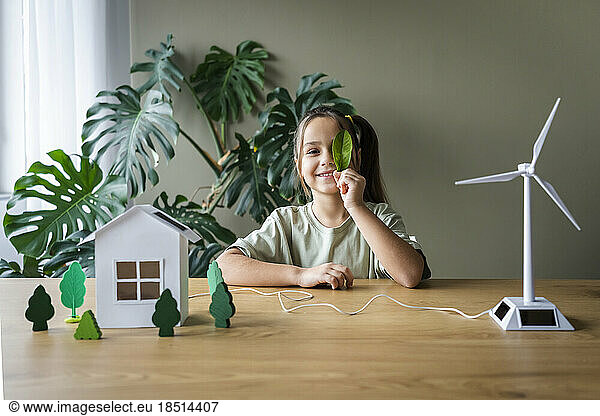 Smiling girl covering eye with leaf sitting at table with wind turbine and house model