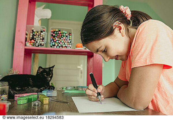 Smiling girl drawing on paper with cat sitting at table