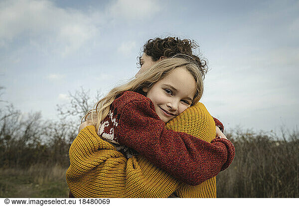 Smiling girl embracing mother in front of sky