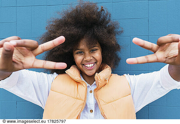 Smiling girl gesturing peace sign in front of blue wall