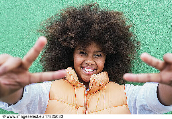 Smiling girl gesturing peace sign in front of wall