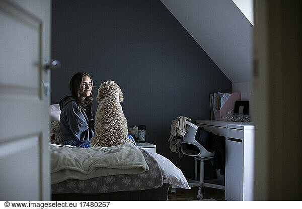 Smiling girl looking at dog while sitting on bed in bedroom