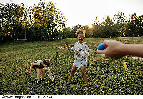 Smiling girl playing with balls while standing by male friend on grass in playground