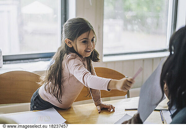 Smiling girl pointing at teacher showing drawing during art class in kindergarten