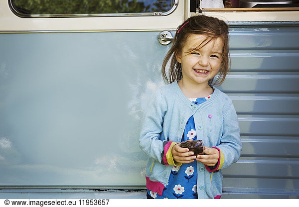 Smiling girl standing in front of blue mobile coffee shop  holding chocolate brownie.
