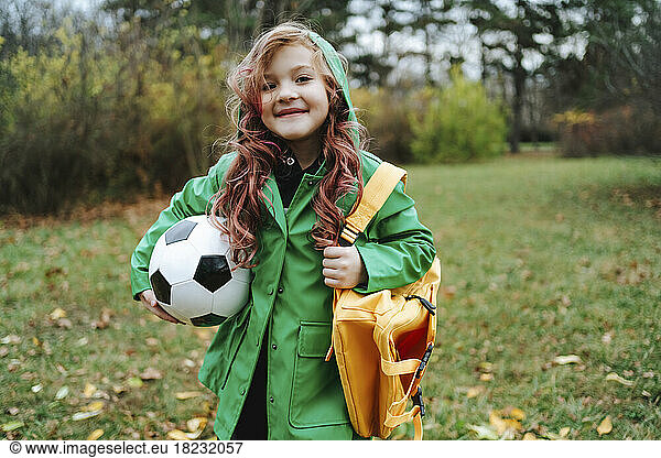Smiling girl standing with backpack and soccer ball at park