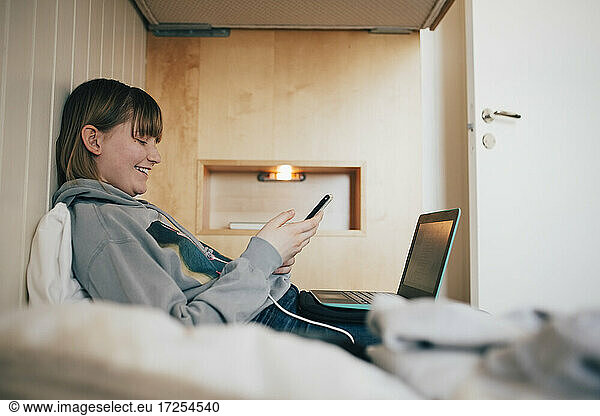 Smiling girl text messaging through mobile phone while sitting on bed at vacation home