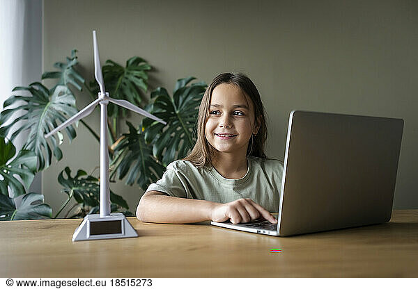 Smiling girl using laptop by wind turbine model on table at home