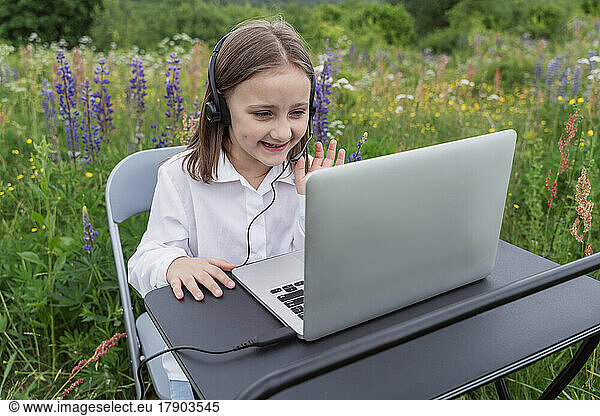 Smiling girl waving at laptop through video call in meadow