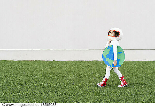 Smiling girl wearing astronaut costume holding earth cut out walking on grass