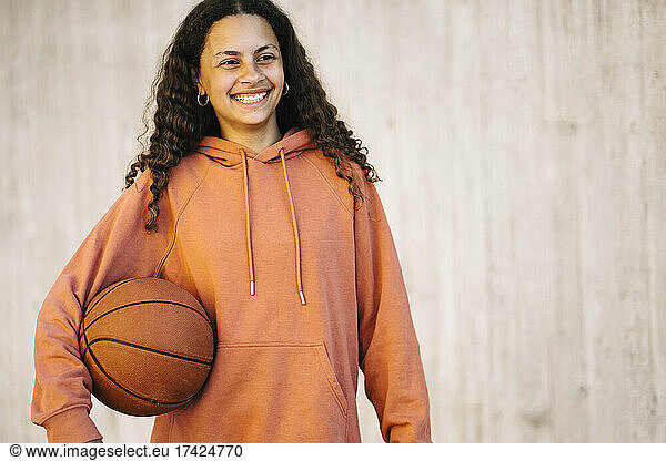 Smiling girl with basketball looking away while standing against wall