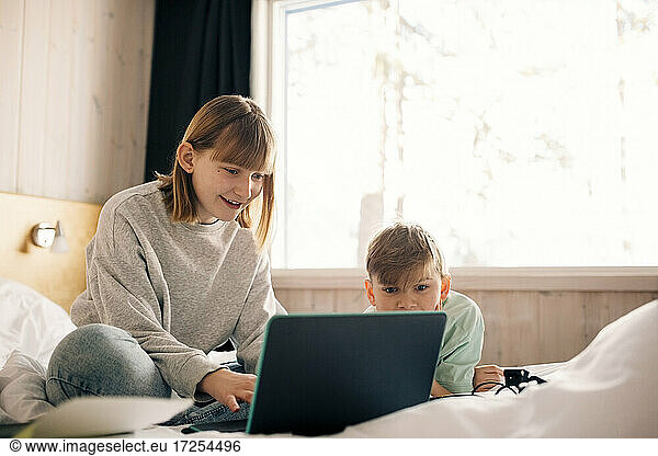 Smiling girl with brother using laptop in bedroom at home