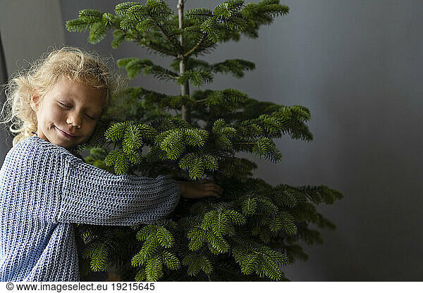 Smiling girl with eyes closed hugging Christmas tree at home
