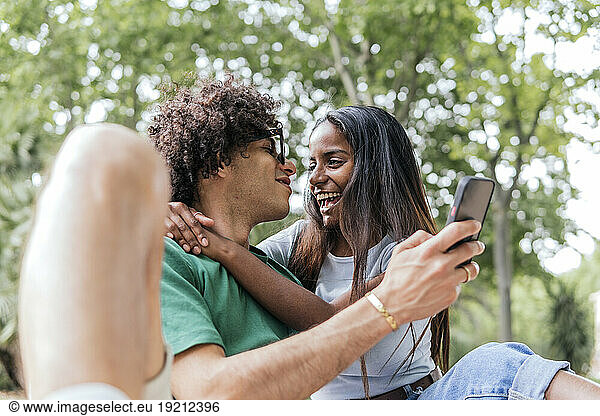 Smiling girlfriend hugging boyfriend with smart phone in park