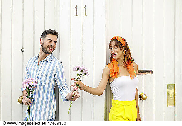 Smiling girlfriend taking flowers while leaning on white door