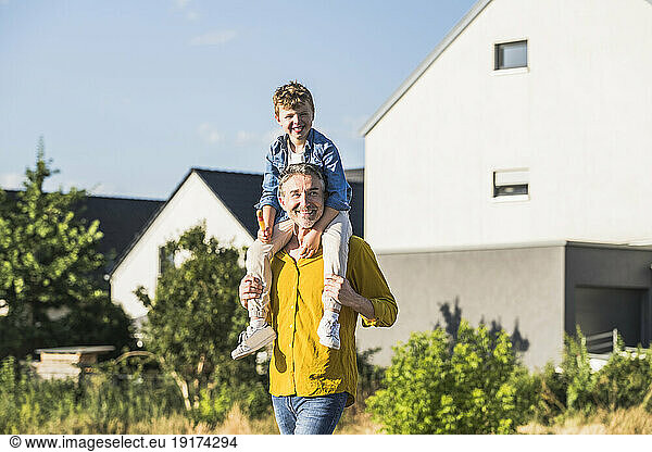 Smiling grandfather carrying grandson on shoulders in front of house
