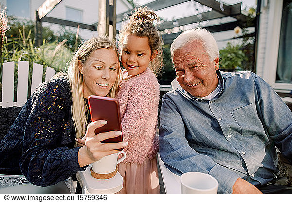 Smiling grandparent and granddaughter taking selfie with mobile phone while sitting in backyard