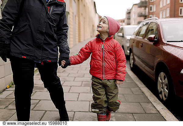 Smiling grandson in warm clothing holding hands of grandmother while walking on footpath