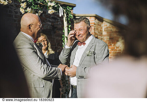 Smiling groom wiping his tears with finger while holding hand of partner by minister on sunny day