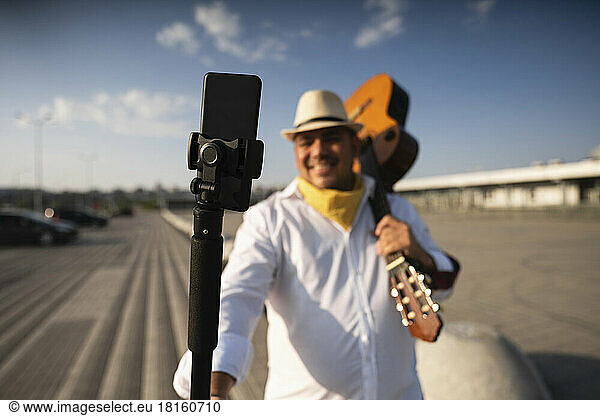 Smiling guitarist holding guitar taking selfie through mobile phone on tripod