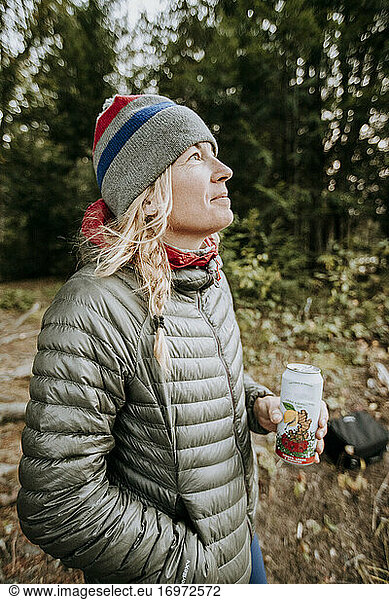 Smiling happy blonde woman in gray jacket and winter hat holding beer.