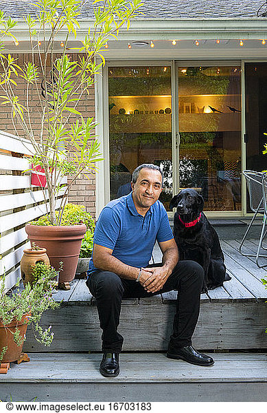 Smiling Hispanic male sit on his deck with black dog