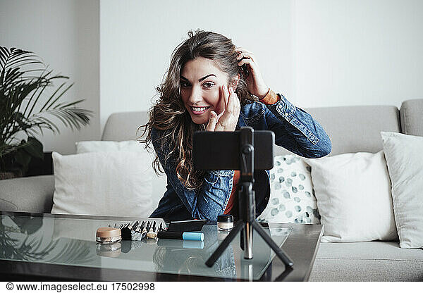 Smiling influencer applying make-up and filming through mobile phone at home studio