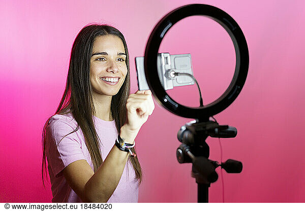 Smiling influencer filming on smart phone in ring light over pink background