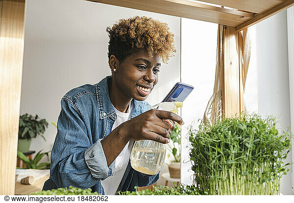 Smiling influencer spraying water on plants and vlogging over mobile phone at home