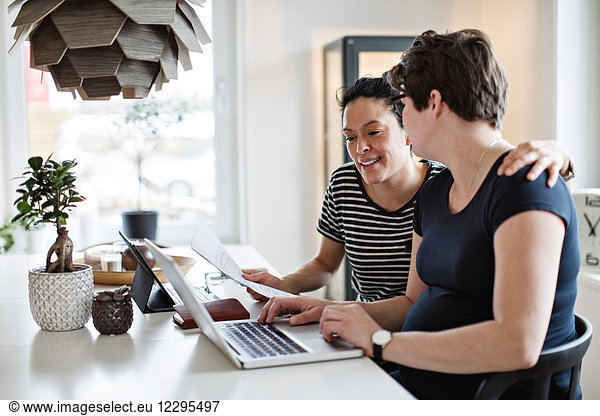 Smiling lesbian couple discussing over bills while using laptop at table