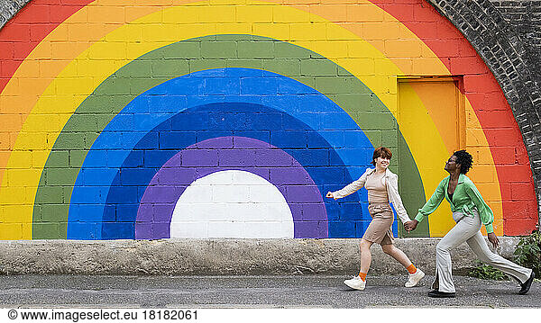 Smiling lesbian couple holding hands running on footpath in front of rainbow colored wall