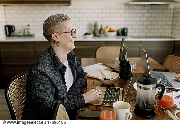 Smiling lesbian professional sitting with laptop at dining table in kitchen