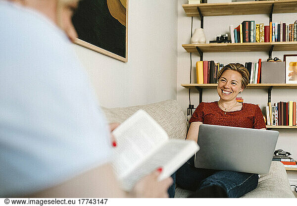 Smiling lesbian woman freelancing on laptop while talking with girlfriend in living room