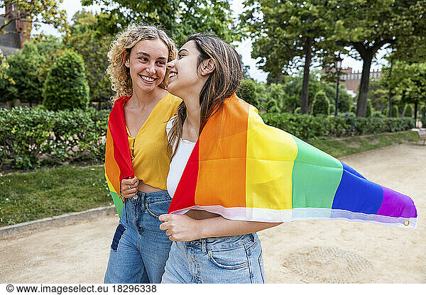 Smiling lesbian young couple covered with rainbow flag walking in park