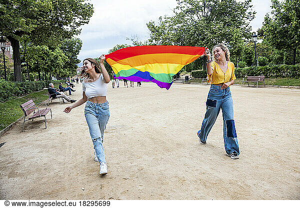Smiling lesbian young couple holding rainbow flag walking in park