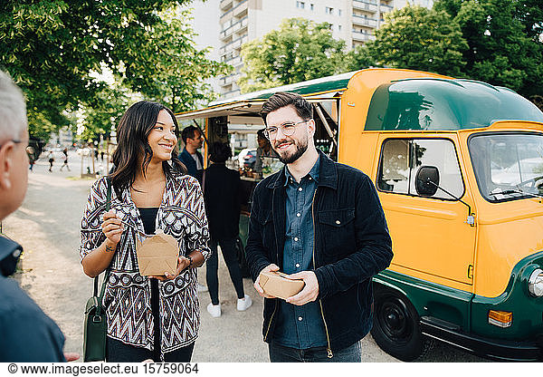 Smiling male and female friend with box standing against food truck