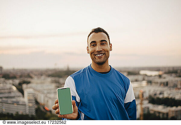 Smiling male athlete holding smart phone against sky during sunset