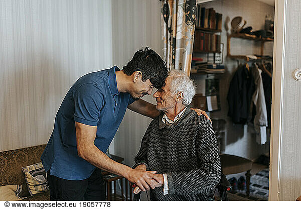 Smiling male care assistant touching forehead with senior man at home