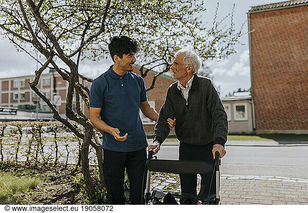Smiling male caregiver assisting senior man with walker on footpath