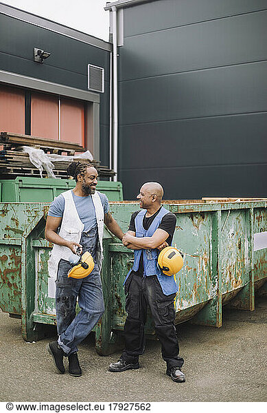 Smiling male colleagues with hardhat looking at each other against against metal container