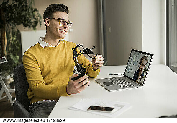 Smiling male engineer with robotic arm attending video conference while sitting at desk
