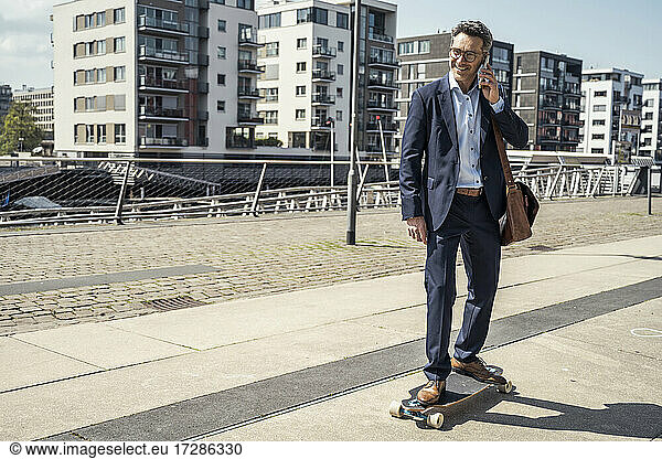 Smiling male entrepreneur talking on mobile phone while skateboarding in city