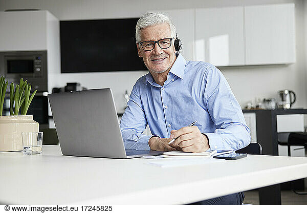 Smiling male entrepreneur with wireless headset and laptop sitting at desk