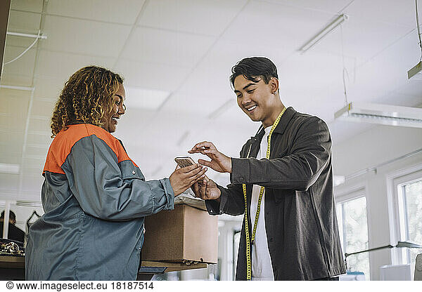 Smiling male fashion designer giving digital signature while receiving package from delivery person at workshop